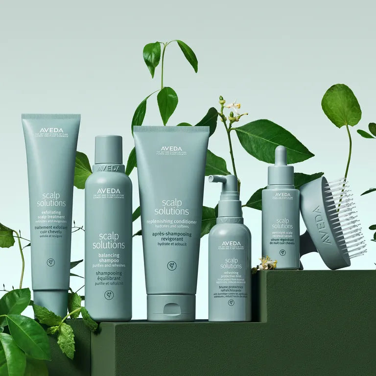 A set of Aveda Scalp Solutions haircare products in muted sage green packaging, arranged on a dark green platform with fresh green leaves against a pale green background.
