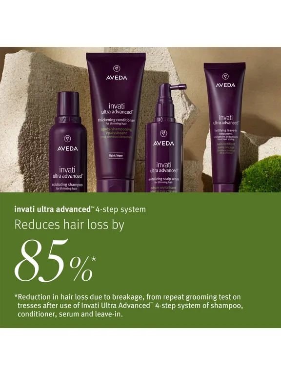Four Aveda Invati Ultra Advanced haircare products in dark purple packaging are displayed against a textured stone background, with a green text banner below featuring promotional copy about reducing hair loss by 85%.