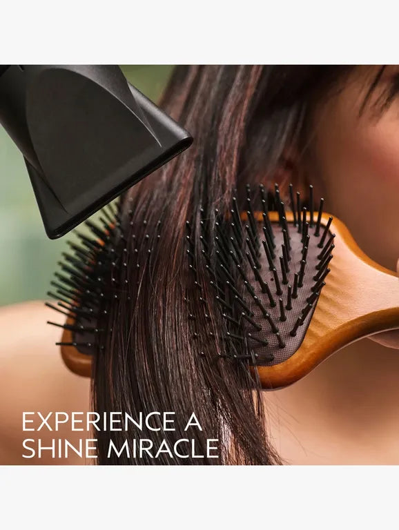 A close-up lifestyle shot of a person using a black hair dryer and a wooden hairbrush on dark brown hair, with the text 'EXPERIENCE A SHINE MIRACLE' at the bottom.