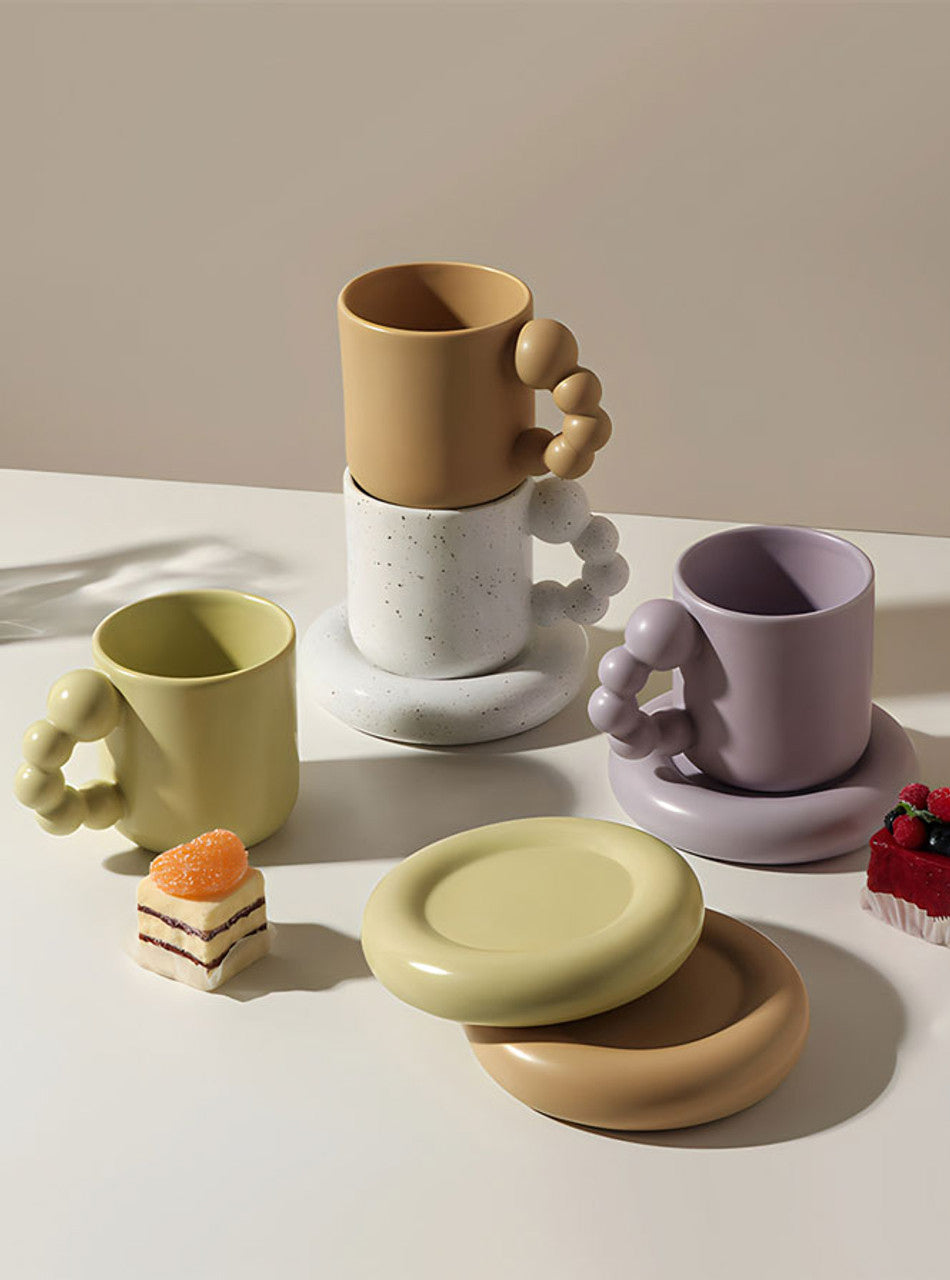 Three ceramic mugs with sculptural bubble handles and matching saucers are arranged on a white tabletop against a warm beige wall. The mugs come in pale yellow-green, tan, and light lavender, with one stacked on top of the other and two standing separately.