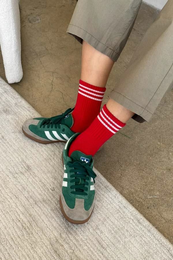 A person wearing red crew socks with white stripes, paired with green and gray sneakers and khaki pants, standing on a textured rug.