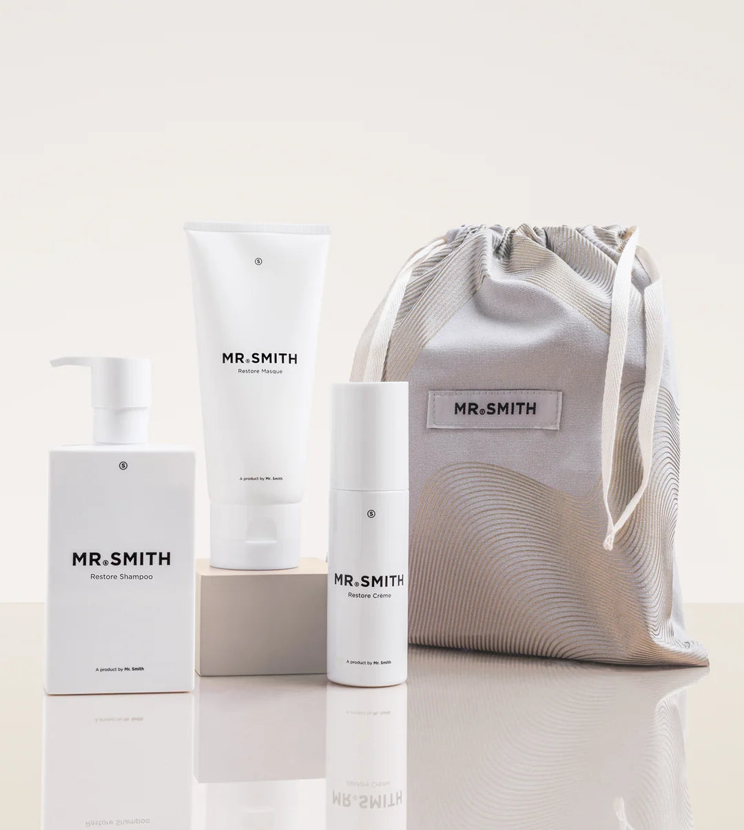 A Mr. Smith haircare gift set featuring a white shampoo bottle on a pedestal, a white conditioner tube, a white cylindrical bottle, and a light gray patterned drawstring pouch, all with the Mr.