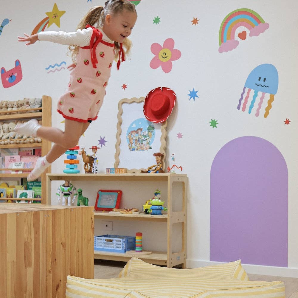 A young girl with blonde hair in a ponytail is jumping excitedly in a colorful children's room. She is wearing a strawberry-print pink dress with red trim.