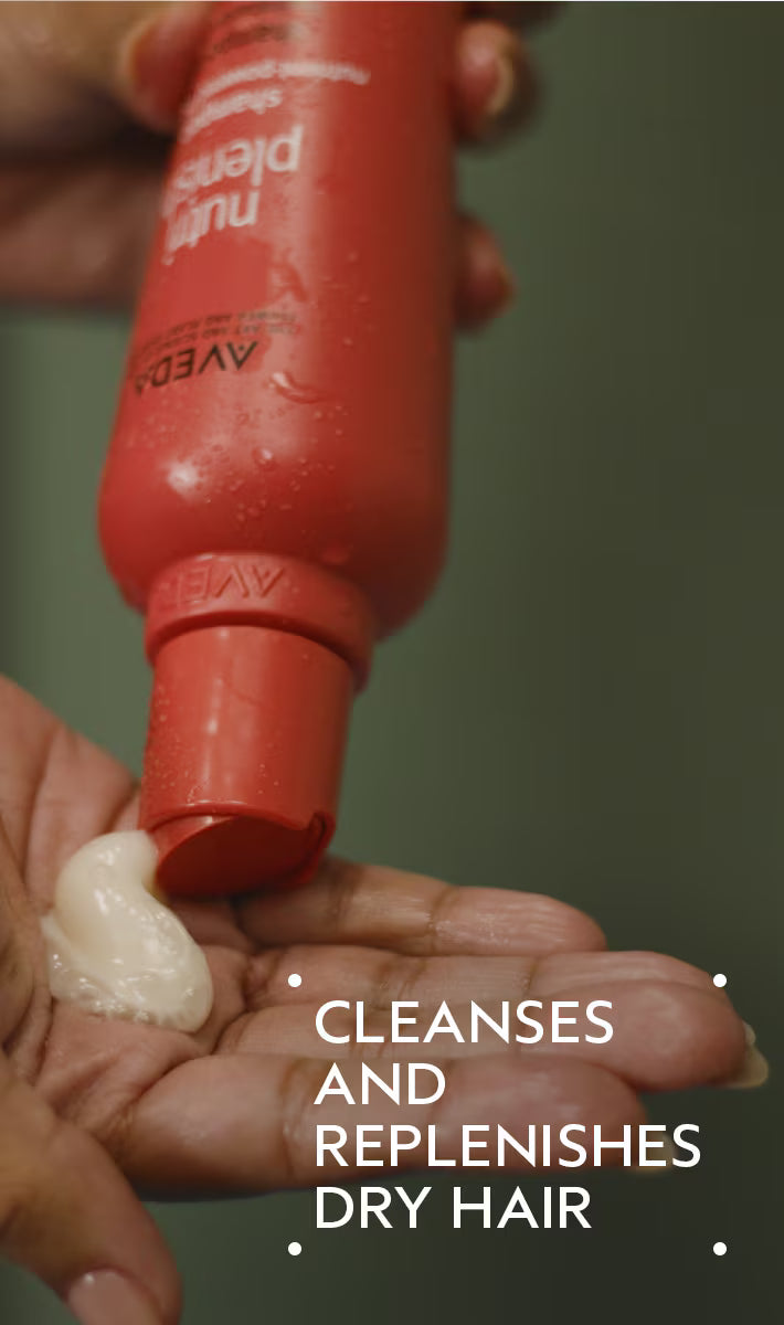 Red bottle of Aveda product being applied to a hand with text 'Cleanses and replenishes dry hair'.