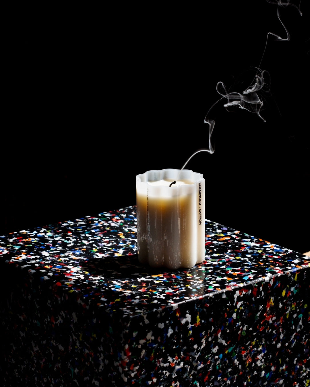 A lit white candle with visible wick and smoke rising, placed on a glossy black speckled cube against a solid black background.