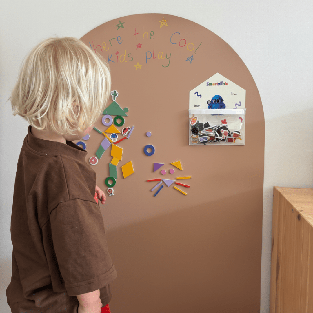 A child with blonde hair wearing a brown shirt stands in front of a large arched brown playboard mounted on a white wall. The playboard has colorful magnetic shapes, a small house-shaped storage pocket with stickers, and the words 'Where the Cool Kids play' written on it.