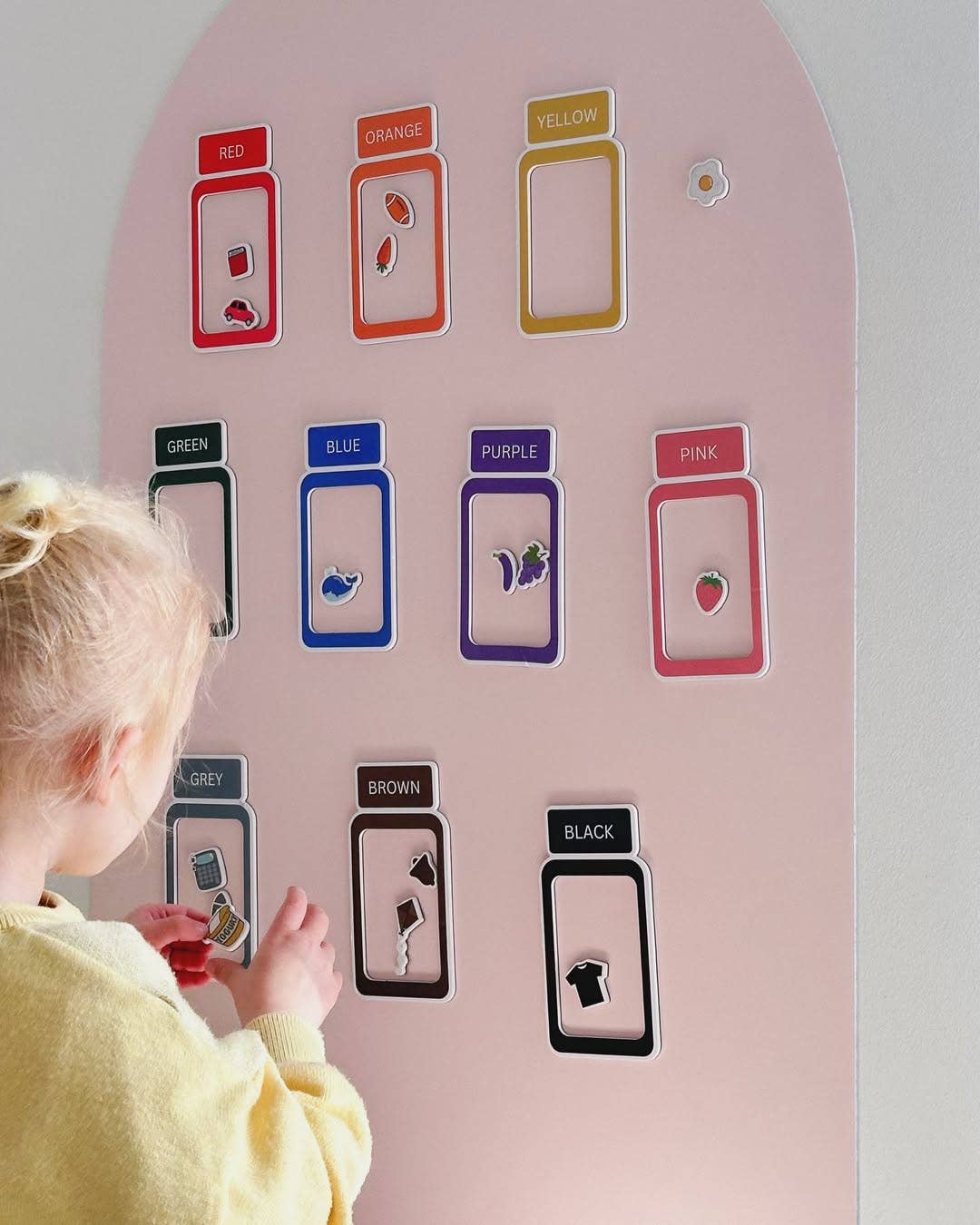 A young child in a yellow sweater is interacting with a pink color learning wall. The wall features labeled color cards (Red, Orange, Yellow, Green, Blue, Purple, Pink, Grey, Brown, Black) with small matching colored objects inside, shown against a light gray wall.