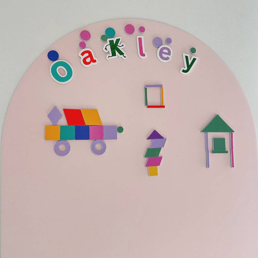 A pastel pink circular wall-mounted playboard with colorful geometric puzzle pieces and the name 'Oakley' spelled out at the top, shown against a plain light wall.