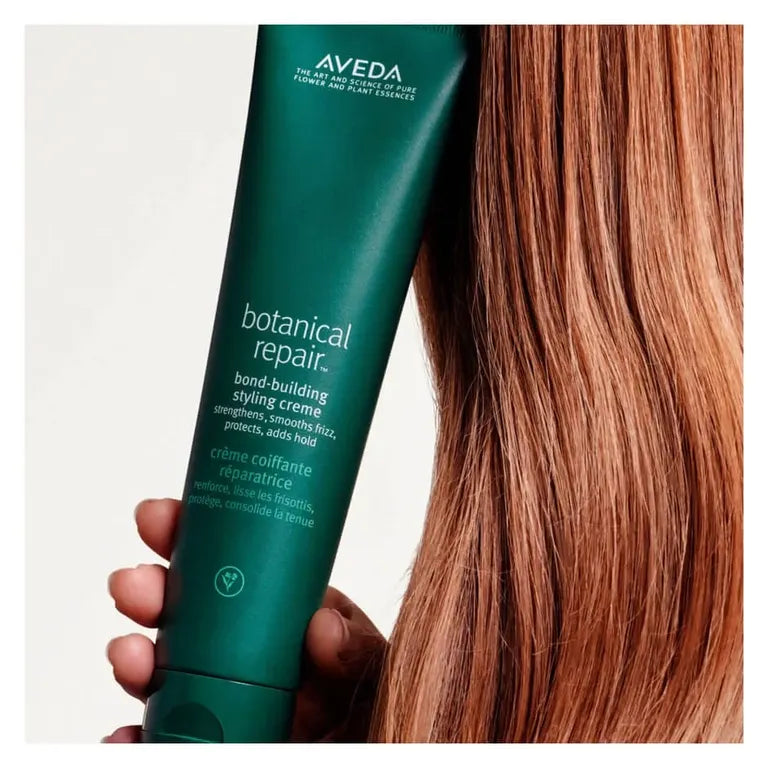 A hand holding a dark green Aveda Botanical Repair bond-building styling creme tube next to long, straight auburn hair on a light gray background.