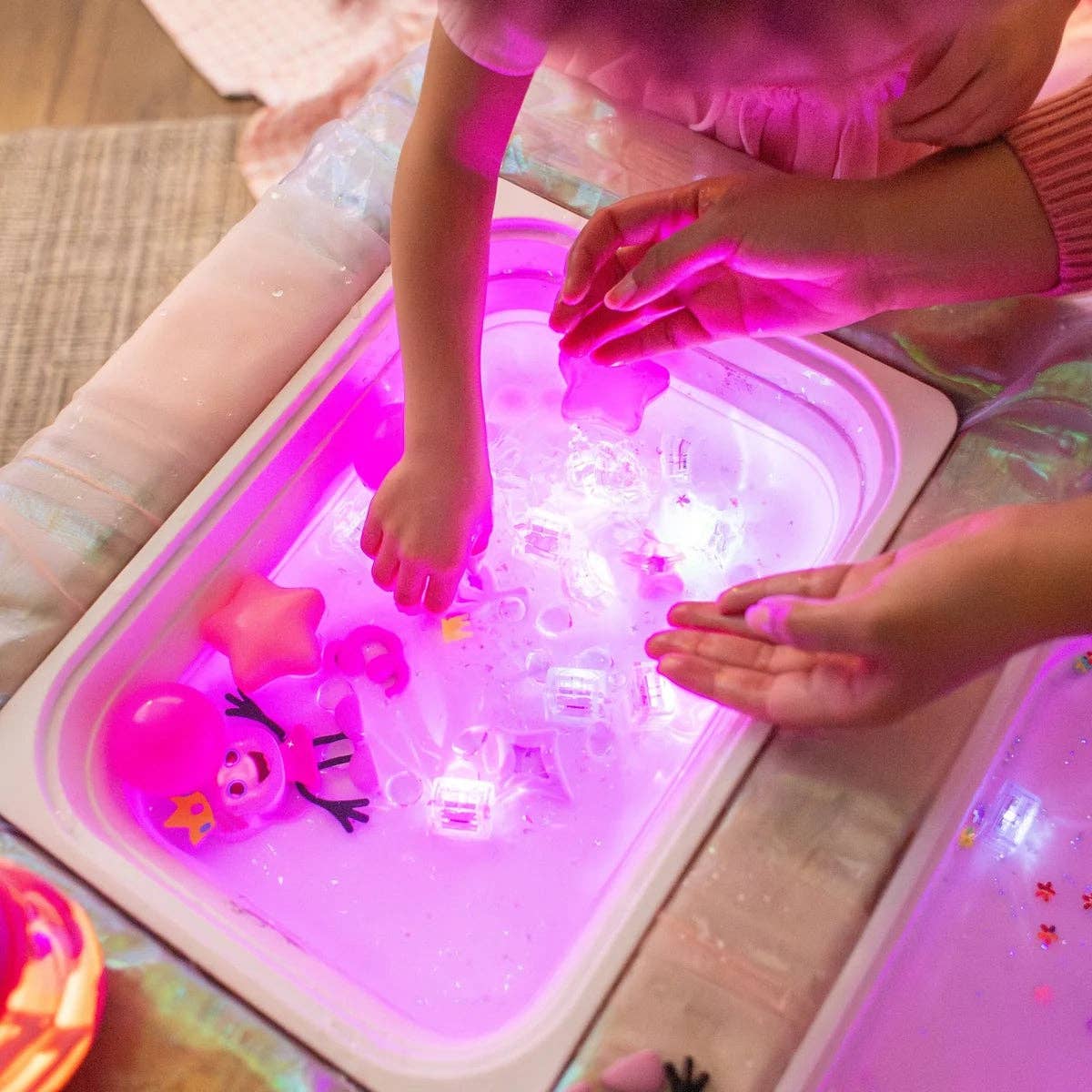 A children's sensory play tray glowing under pink LED lights, filled with water, ice, and colorful toys, with a child's hands reaching into the tray.