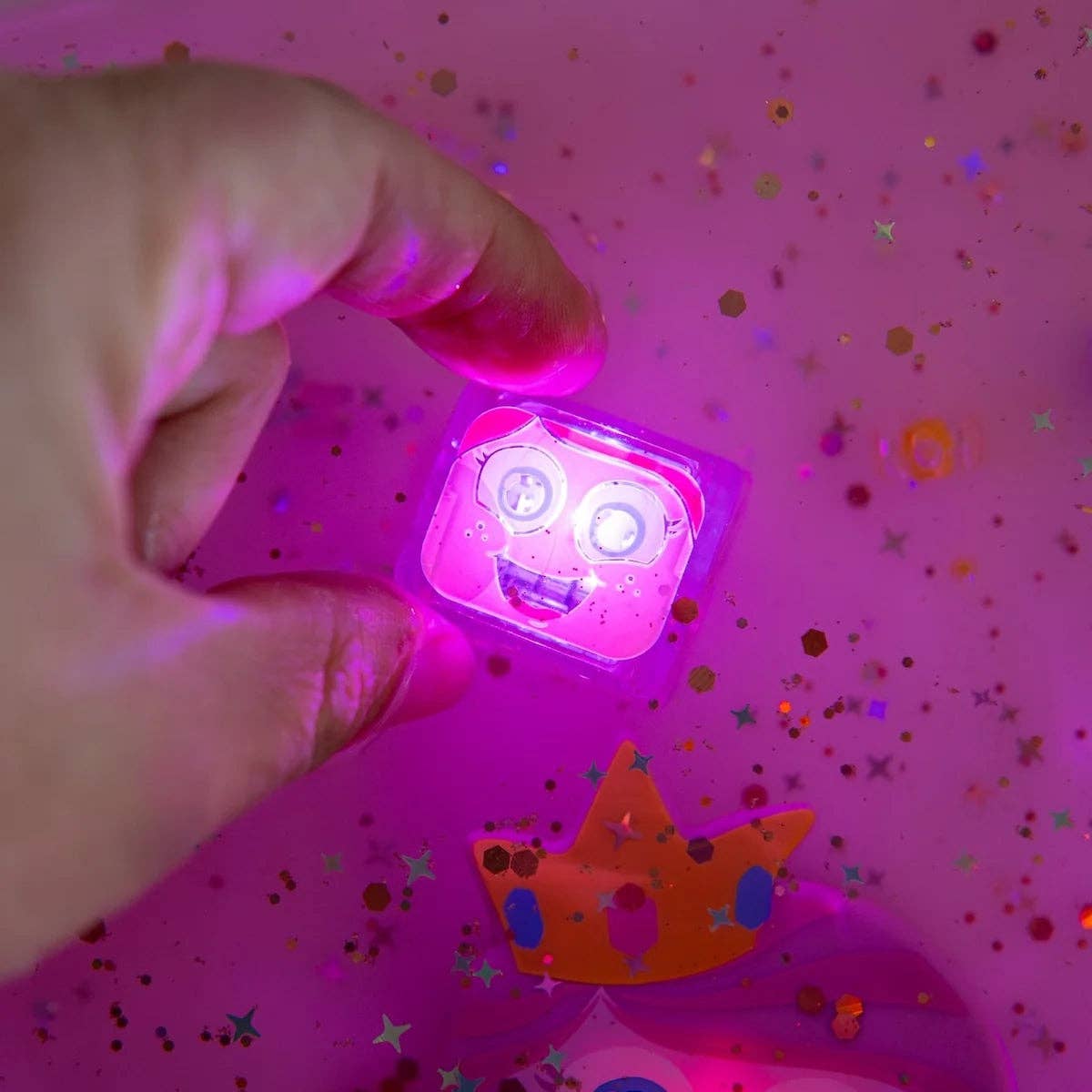 Hand holding a glowing pink object with a face on a pink background with confetti