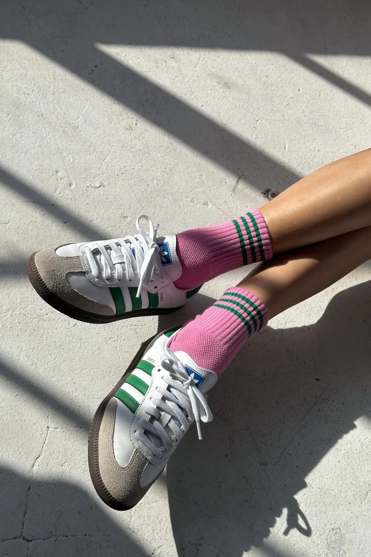 A person's legs crossed, wearing pink crew socks with green and black stripes, paired with white and gray Adidas Samba sneakers on a concrete floor with diagonal shadows.